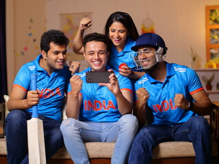 A group of fans in Indian jerseys sitting on couch and watching live cricket match on phone -...