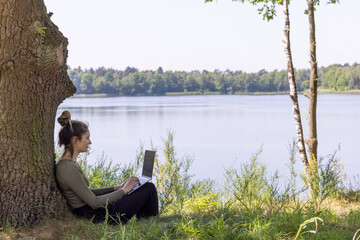 Embrace the freedom of remote freelance work with this captivating scene. A young woman, a dedicated freelancer, effortlessly balances her work and nature's allure. She sits by a tranquil forest lake