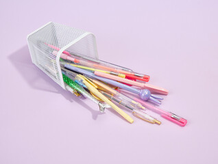 Various colored pens fall out of the stationery stand on a purple background. School supplies composition.