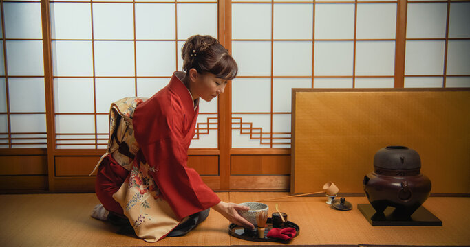 Adult Japanese Female Preparing Matcha Green Tea At Home While Sitting On A Tatami Floor. Asian Woman In Traditional Red Kimono With FLower Ornaments Trying The Tea From A Bowl