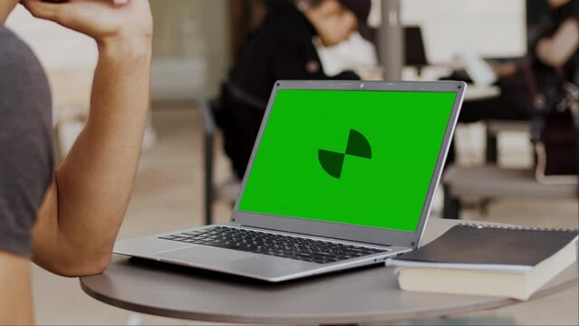 Young man sits at a table in a cafe at a laptop with a mockup green screen and nods affirmatively close up. Concept of modern remote work with flexible schedule