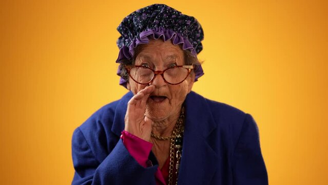 Funny portrait of smiling happy crazy toothless grandmother with wrinkled skin puts hand to mouth to tell a secret isolated on yellow background studio
