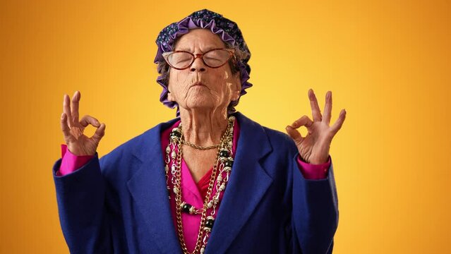 Funny crazy grandmother senior woman saying OM, 80s, meditating doing yoga exercise isolated on yellow background. Concept of youthful old female.