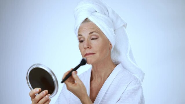 Happy senior european woman applying foundation powder concealer with brush, smiling, female doing makeup wearing bathrobe and towel on head with copy space