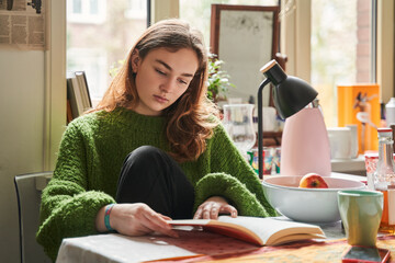 Calm teen girl reading book while sitting at kitchen during breakfast