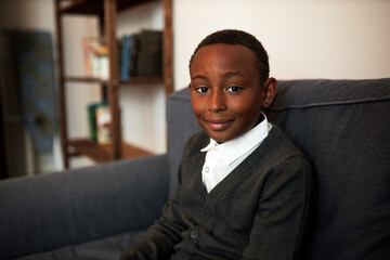 Horizontal portrait of cute little black schoolboy sitting on sofa at home in white shirt and...