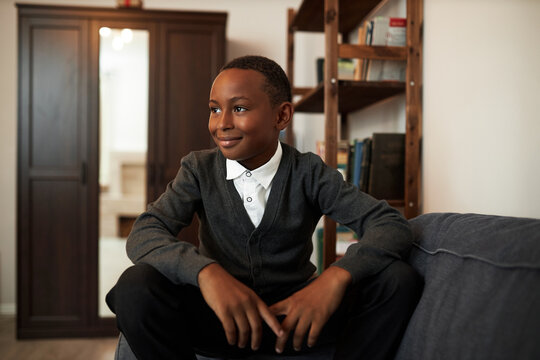 Cool african american male schoolkid in uniform having rest on couch in classroom during break, looking aside with smiling facial expression sitting on armrest putting hands on knees