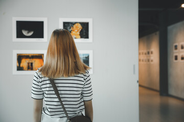 Fototapeta na wymiar Asian young woman standing she looking art gallery in front of colorful framed paintings pictures on white wall, female watch at photo frame to leaning against at exhibit museum, Back view