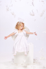 Fototapeta premium Happy, smiling, positive little girl, child in costume of angel sitting isolated over white studio background with flying feathers. Concept of childhood, imagination, fantasy, holidays, holidays