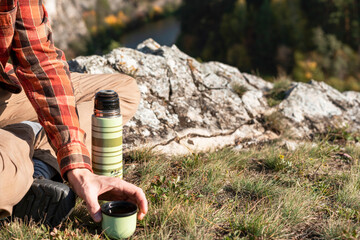 Fototapeta premium unrecognizable man tourist in red shirt drinking tea from thermos while resting on rocks by river in fall hiking and relaxing in nature