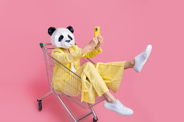 Full body photo of weird 3d panda mask lady sit inside store pushcart hold smart phone make selfie isolated on pink color background © deagreez
