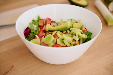 Still life with a white bowl of fresh salad with organic vegetables, avocado fruit and greens on a wooden table