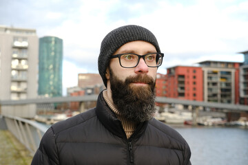 young bearded man in coat and hat stands on shore river Main, modern buildings in Frankfurt, skyscraper Westhafen Tower in Gutleitviertel district, concept of winter city walks, tourism