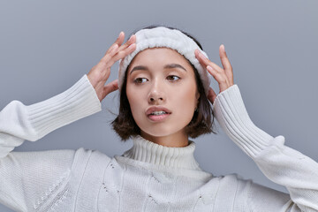 stylish asian woman in knitted sweater adjusting warm headband on grey, winter fashion photography