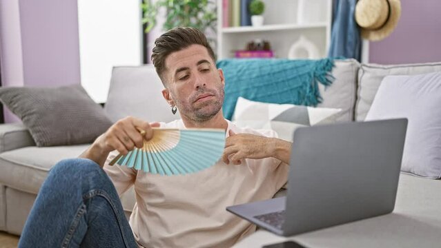 Hot problem! young, focused hispanic man sweating while sitting on living room floor using handfan at home, working online on laptop.
