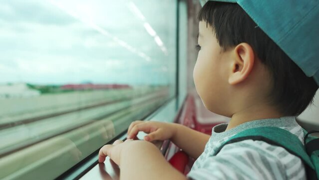 Little Asian Japanese boy in backpack and blue cap looking through the window while going to the school by commuter train.