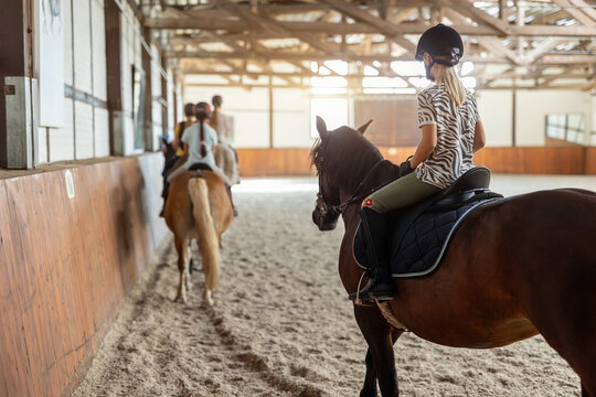 Horse riding school. Little children girls at group training equestrian lessons in indoor ranch horse riding hall. Cute little beginner blond girl kid in helmet sitting on brown horse horseback