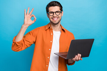 Photo of positive handsome guy hold netbook demonstrate okey symbol eshopping isolated on blue...