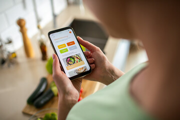 Close-up of a young woman looking for recipes on her smartphone while preparing a organic meal in the kitchen - obrazy, fototapety, plakaty