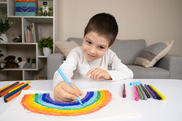 A small child draws a rainbow with felt-tip pens in a sketchbook while sitting at a table