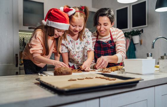 Cute little girls in red Santa hats with mother making homemade dough Christmas gingerbread cookies using cookie cutters together in home kitchen. Happy family holidays preparation, childhood concept.