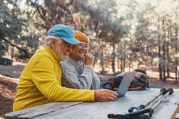 Head shot portrait close up of cute couple of old middle age people using computer pc outdoors sitting at a wooden table in the forest of mountain in nature with trees around them..