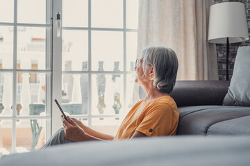 Smiling middle-aged Caucasian woman sit on couch in living room browsing wireless Internet on tablet, happy modern senior female relax on ground at home using pad device, elderly technology concept.
