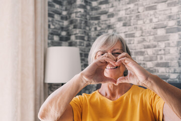 Elderly woman sit in living room connected fingers showing heart symbol close up, old people...