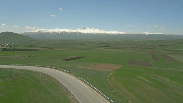Drone video. Straight fly-by along a green stony hill, a highway with a passing van, a small village and the high snowy mountain of Aragats. Sunny weather, blue sky with clouds. Lori region, Armenia.