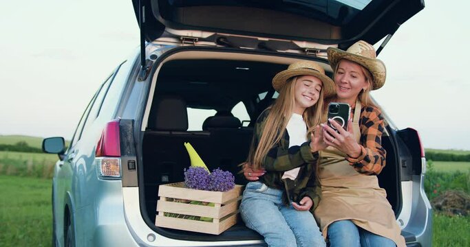 Family of farmers, woman and girl in aprons and hats siting in trunk of car next to a wooden box with fresh flowers using smartphone in field at sunset. Small family business concept. Slow motion.