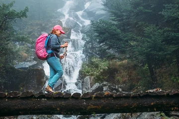 Crédence de cuisine en verre imprimé Makalu Young woman with backpack and trekking poles crossing wooden bridge near power mountain river waterfall during Makalu Barun National Park trek in Nepal. Mountain hiking and active people concept image