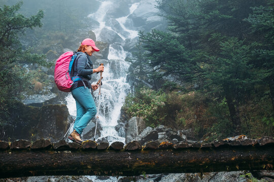 Young woman with backpack and trekking poles crossing wooden bridge near power mountain river waterfall during Makalu Barun National Park trek in Nepal. Mountain hiking and active people concept image
