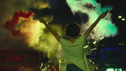 Young woman standing in middle of sport cars and waving , holding colorful smoke bombs flares . Girl starting muscle car drag racing at night . Back view slow motion . Ready for racing . Crowd of fans