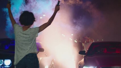 Young woman standing in middle of sport cars and waving , holding colorful smoke bombs flares . Girl starting muscle car drag racing at night . Back view slow motion . Ready for racing . Crowd of fans