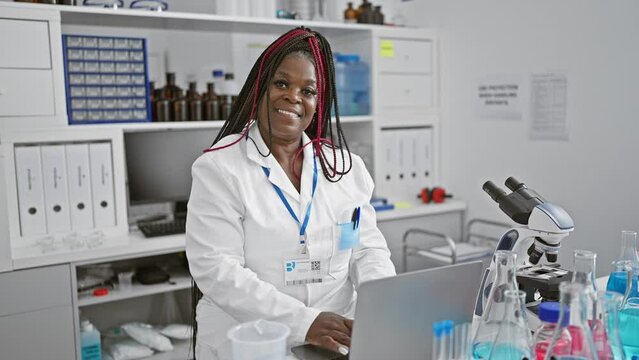 Confident african american woman, smiling widely, fully immersed in scientific research on her laptop in the bustling medical lab, beautifully braided hair catching the light as she works.
