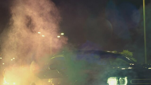 Young woman standing in middle of sport cars and waving , holding colorful smoke bombs flares . Girl starting muscle car drag racing at night . Back view slow motion . Ready for racing . Crowd of fans