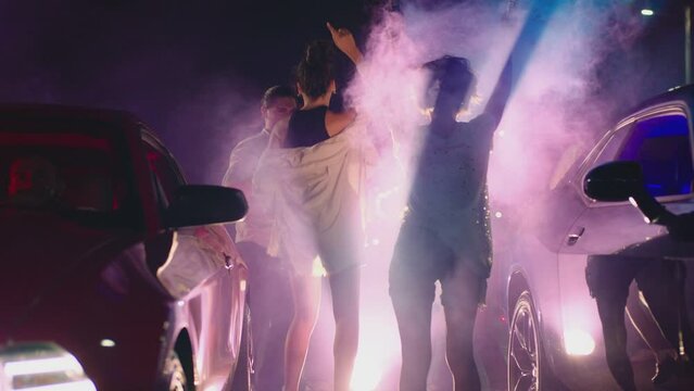Young woman standing in middle of sport cars and waving , holding colorful smoke bombs flares . Girl starting muscle car drag racing at night . Back view slow motion . Ready for racing . Crowd of fans