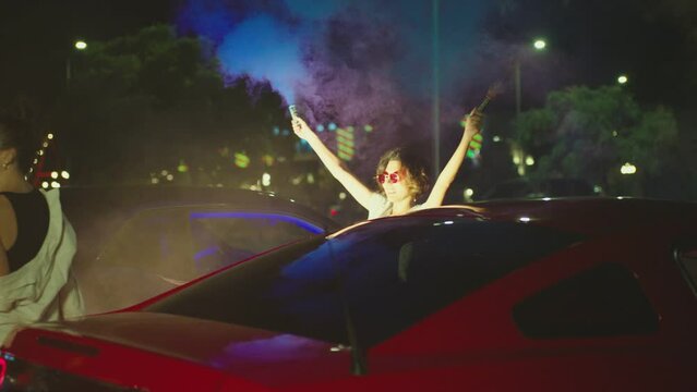 Young woman standing in middle of sport cars and waving , holding colorful smoke bombs flares . Girl starting muscle car drag racing at night . Back view slow motion . Ready for racing . Crowd of fans
