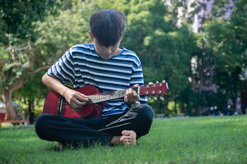 Obraz premium Asian cute boy playing guitar in the front yard of his own house happily, soft focus, concept for recreational activity and free times hobbies of children around the world.