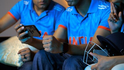 Close-up pan shot of friends deeply engrossed in a crucial cricket match displayed on their smartphone Indian jersey. Indian cricket fans watching a cricket match watching World Cup at home cri... - Powered by Adobe