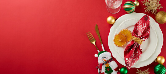 Elevate your Christmas dining experience. Top view of plates, cutlery, glass, snowflakes, xmas balls, snowman, dry orange slices on red background with advert space
