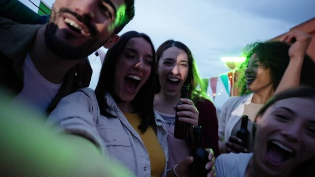 Young Caucasian woman excited shooting selfie video phone at party. Attractive female looking cheerful on camera friends dancing outdoors at sunset. People gathered on rooftop celebrating birthday.