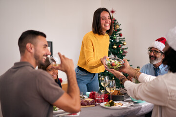 Beautiful young smiling Caucasian woman standing passing salad at Thanksgiving meal. Happiness at December festive vacation. Multi-generational people enjoying family reunion together Christmas. - obrazy, fototapety, plakaty