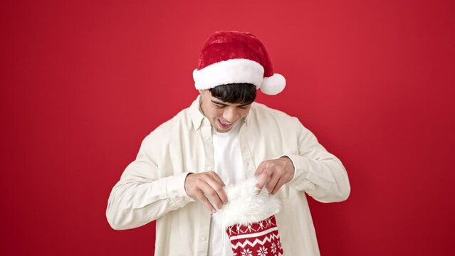 Young hispanic man wearing christmas hat looking inside of sock over isolated red background