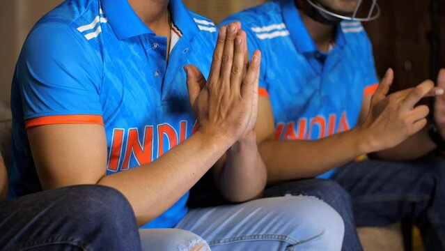 Close-up of a boy praying for the Indian cricket team to win, praying with folded hands, wearing an Indian cricket team jersey. Three young Indian cricket fans are watching a cricket match at home