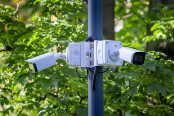 Two outdoor city video security cameras on a pole on background of green leaves. Safe and comfortable urban environment. Control and combating crime. Street security, safety and city life improvement
