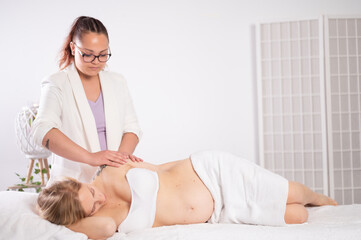 Ethnic masseuse massaging resting young pregnant woman on bed in parlor