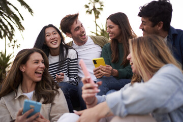 Group of diverse people enjoying day together outdoors with mobiles in hand. Young multiracial friends using cell phone. Beautiful millennial sitting together laughing excited at college class break.