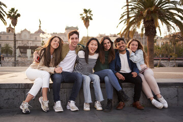 Portrait of joyful group of happy young multi-ethnic friends enjoying vacation in European city. Multiracial people sitting in line in square looking at camera for souvenir photo of trip abroad. 
