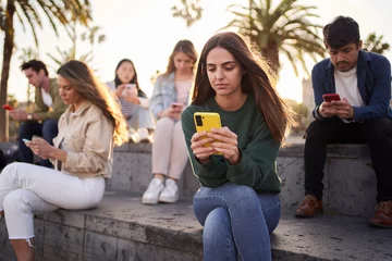 Foto op Canvas Young Caucasian girl sitting concentrated typing on mobile with group friends in background. Serious beautiful woman addicted to technologies using phone outdoors. Social isolation new generations. © CarlosBarquero
